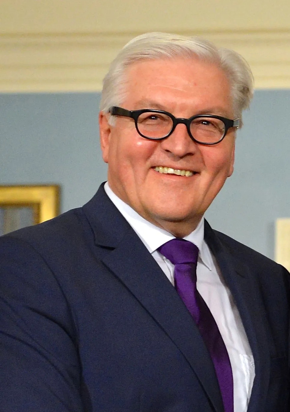 Frank-Walter Steinmeier smiling, wearing dark-rimmed glasses, a navy suit and a purple tie